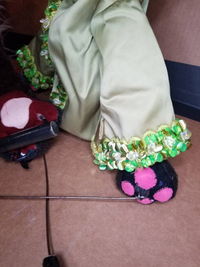 May include: A green and gold puppet with a black and pink foot. The puppet is wearing a green and gold costume with sequins. The puppet is sitting on a brown surface.