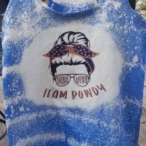 May include: A blue tank top with a bleach-splattered design. The tank top features a graphic of a woman with a bun, wearing sunglasses and a bandana. The text "VISA VISA TEAM ROWDY" is printed on the tank top.