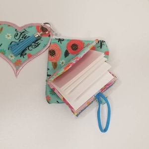 May include: A small, floral patterned notebook with a blue tassel keychain. The notebook is open to reveal blank pages and has a blue elastic closure.