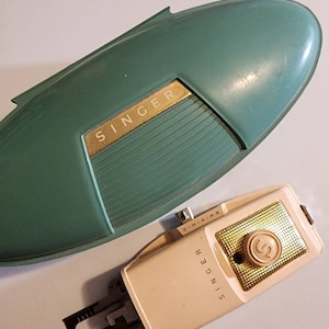 May include: A vintage Singer sewing machine foot pedal with a green case. The foot pedal is beige with gold accents and the word "SINGER" printed on it. The case is a light green oval shape with the word "SINGER" printed in gold letters.