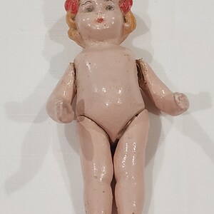 May include: A small, porcelain doll with blonde hair and red bows. The doll is wearing a pink dress and has a painted face.