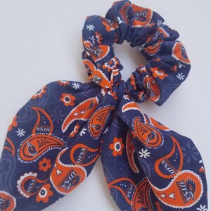 May include: A blue and orange paisley patterned hair scrunchie with a bow. The scrunchie has the text "UTSA" repeated throughout the pattern.
