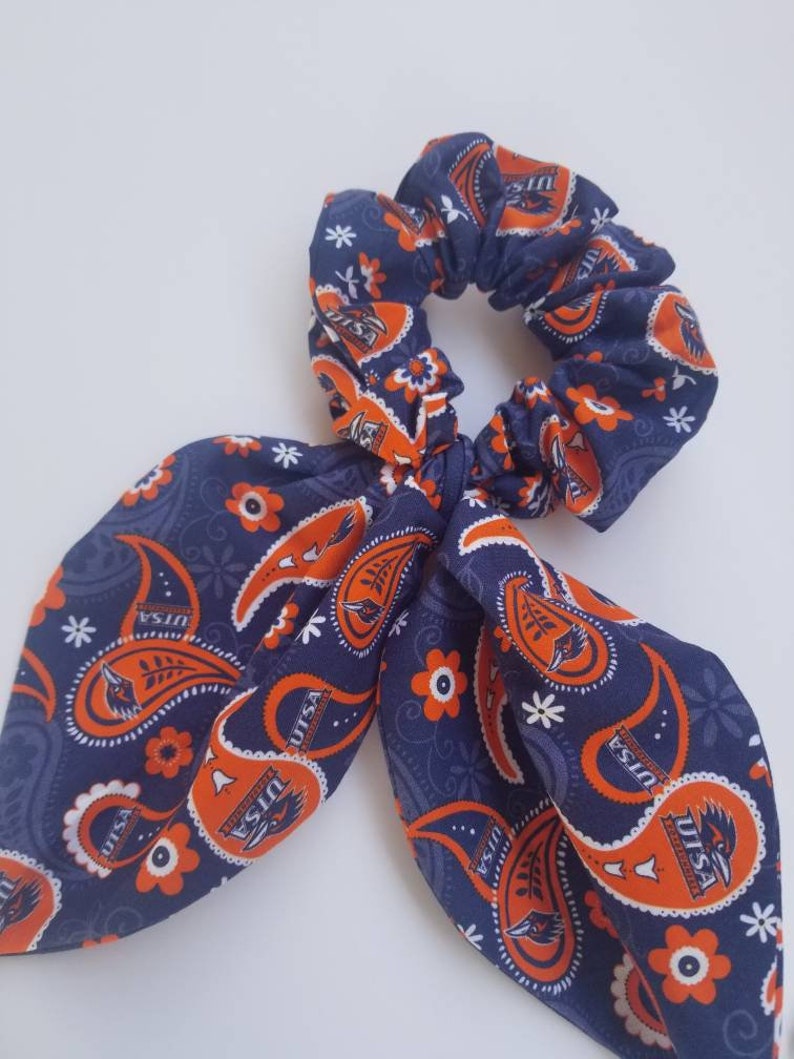 May include: A blue and orange paisley patterned hair scrunchie with a bow. The scrunchie has the text "UTSA" repeated throughout the pattern.