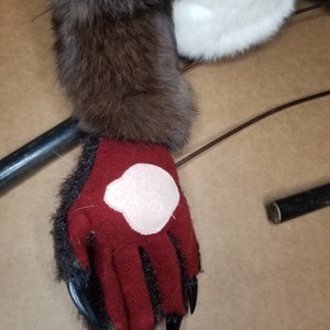 May include: A close-up of a red furry hand with black claws. The hand is wearing a red glove with a pink patch on the back.