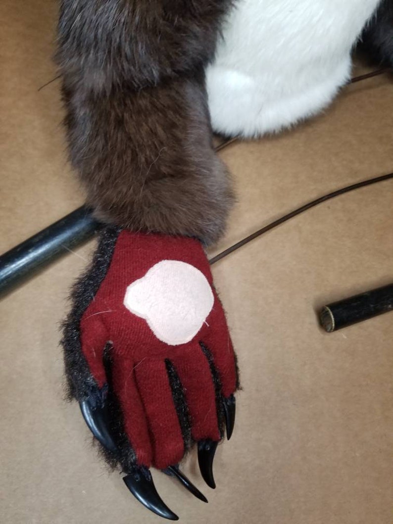 May include: A close-up of a red furry hand with black claws. The hand is wearing a red glove with a pink patch on the back.