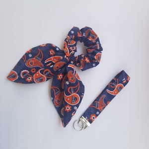 May include: A blue and orange paisley patterned hair scrunchie and matching key chain. The scrunchie is made of fabric and has a gathered elastic band. The key chain has a silver key ring.