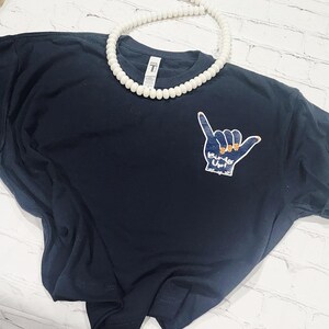 May include: Navy blue t-shirt with a hand gesture graphic that says "Birds Up!". The graphic is in blue and yellow. A white beaded necklace is draped across the shirt.