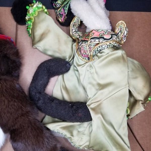 May include: A green, gold, and pink puppet with a black and white face. The puppet is wearing a green satin robe with gold trim and a pink and green patterned scarf. The puppet's arms are outstretched and its legs are crossed.