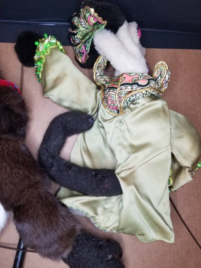 May include: A green, gold, and pink puppet with a black and white face. The puppet is wearing a green satin robe with gold trim and a pink and green patterned scarf. The puppet's arms are outstretched and its legs are crossed.