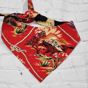 May include: Red bandana with a cowboy riding a horse on a desert landscape. The bandana has a white rope design around the edges.