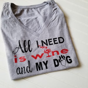 May include: A heather gray V-neck t-shirt with the text "All I need is wine and my dog." The text is in black and red, with a wine glass graphic and a paw print with a heart.