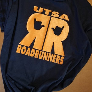 May include: A navy blue t-shirt with an orange graphic of the letters "UTSA" and "RR" with a hand gesture above each letter. The text "ROADRUNNERS" is below the letters.