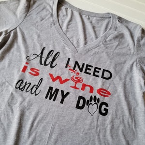 May include: A heather gray V-neck t-shirt with the text "All I need is wine and my dog." The words "wine" and "dog" are in red and black, with a wine glass and paw print graphic.