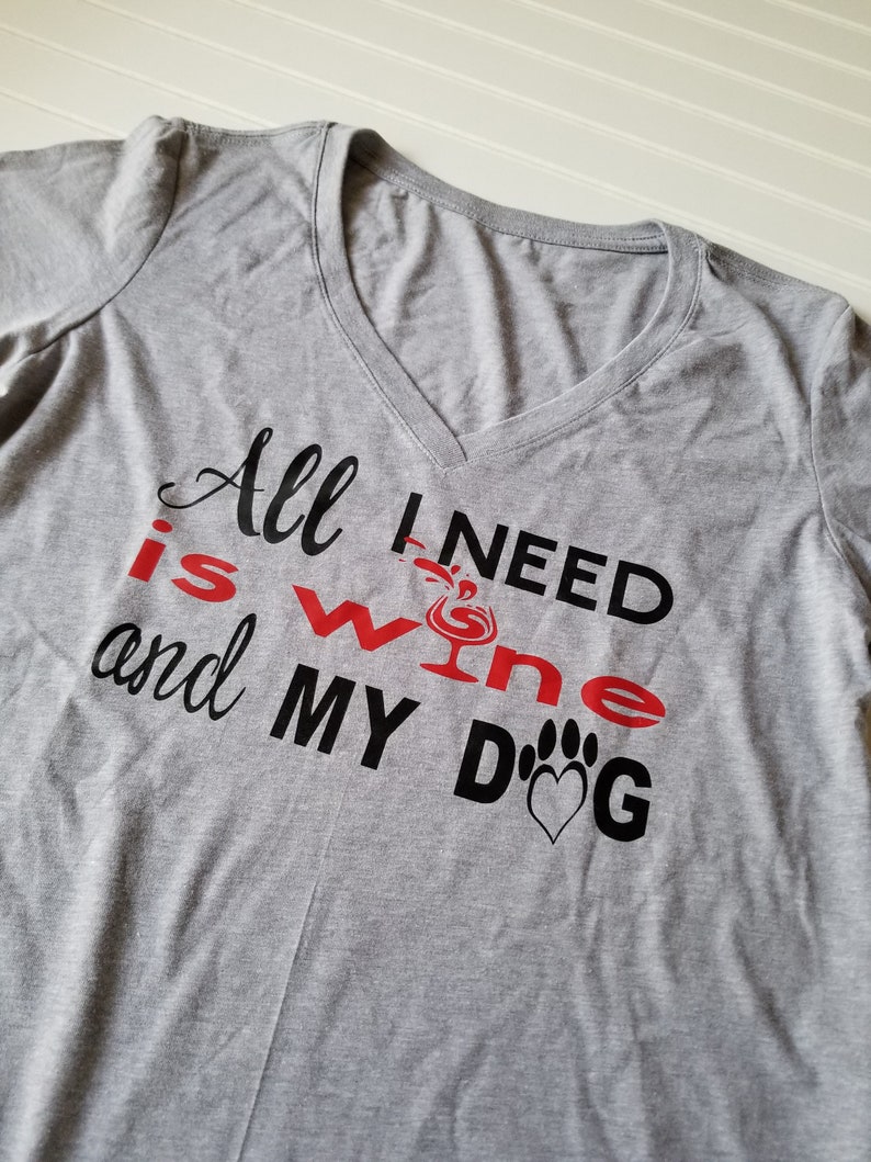 May include: A heather gray V-neck t-shirt with the text "All I need is wine and my dog." The words "wine" and "dog" are in red and black, with a wine glass and paw print graphic.