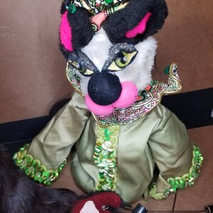 May include: A hand puppet with a black and white face, pink nose, and green eyes. The puppet is wearing a green and gold sequined robe with a pink and green patterned collar. The puppet is also wearing a black and gold crown with green jewels.