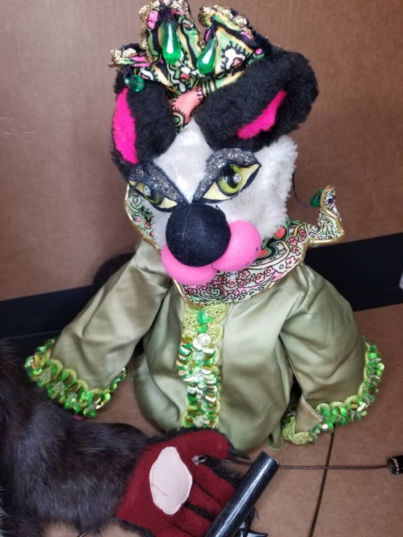 May include: A hand puppet with a black and white face, pink nose, and green eyes. The puppet is wearing a green and gold sequined robe with a pink and green patterned collar. The puppet is also wearing a black and gold crown with green jewels.