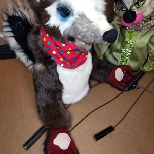 May include: Two hand puppets, one is a gray and white wolf with a red and white polka dot scarf, the other is a green and black cat with a pink nose and a green and gold headpiece. Both puppets have red gloves and are attached to strings.