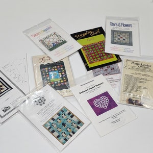 May include: A collection of quilting patterns featuring various designs, including apples, stars, flowers, penguins, and a heart. The patterns are printed on white paper with colorful illustrations and text. The patterns are packaged in clear plastic sleeves.