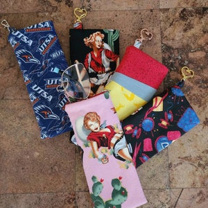 May include: Six different eyeglass cases with various designs. The cases are made of fabric and have a zipper closure. The designs include a blue and white UTSA logo, a red and white pin-up girl, a yellow and blue geometric pattern, a black and white sewing themed pattern, and a pink and green cactus pattern.