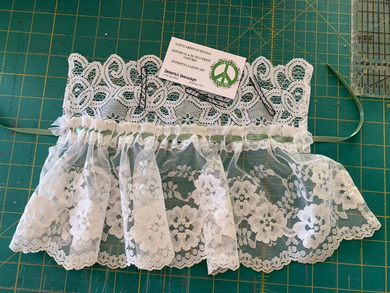 Puede incluir: Borde de encaje blanco con una cinta verde y una tarjeta de visita que dice "Native American Regalia, Historical Re-enactment,  Kythera Garden Art, Deborah Jean Designs".