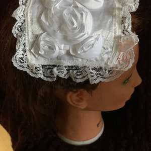May include: White satin headband with a large white satin rose and smaller white satin roses. The headband is trimmed with white lace.