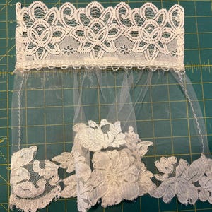 May include: A white lace trim featuring a wide, intricate floral pattern at the top edge, connected to a sheer tulle veil section. The bottom edge is adorned with a dense, scalloped floral lace applique, creating a decorative border.