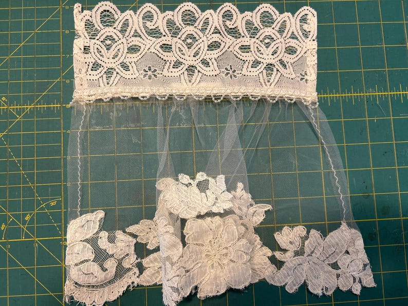 May include: A white lace trim featuring a wide, intricate floral pattern at the top edge, connected to a sheer tulle veil section. The bottom edge is adorned with a dense, scalloped floral lace applique, creating a decorative border.