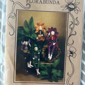May include: A package featuring "Barbara Willis Designs FLORABUNDA" with illustrations of seated dolls. The dolls are dressed in floral-themed costumes, including a sunflower, pansy, and petunia. The package is brown with decorative floral accents.