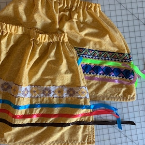 May include: Two yellow fabric skirts with colorful ribbon trim. The top skirt has a green, blue, and pink ribbon trim. The bottom skirt has a blue, red, and brown ribbon trim.