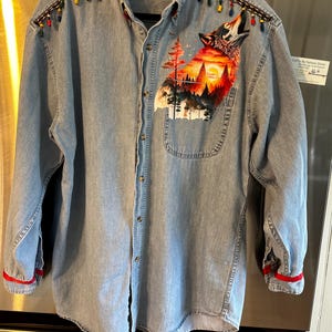 May include: A light blue denim button-down shirt featuring a graphic print of a howling wolf against a sunset forest landscape on the chest. The shoulders are accented with dark trim and colorful beads, while the collar and cuffs feature red trim.