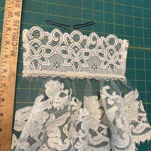 May include: A white lace hair accessory featuring a wide floral lace band with a scalloped edge, attached to a sheer tulle veil with floral lace appliques. Two black metal bobby pins are placed above the lace. A wooden ruler shows a length of 6 inches.