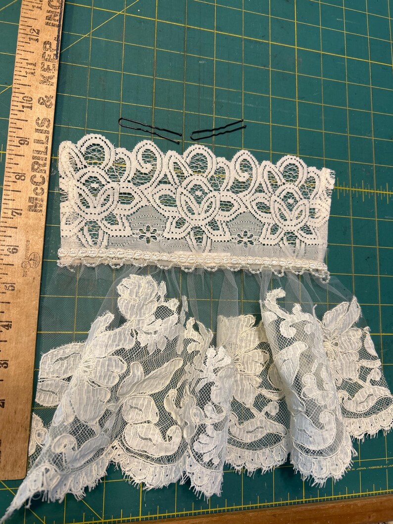 May include: A white lace hair accessory featuring a wide floral lace band with a scalloped edge, attached to a sheer tulle veil with floral lace appliques. Two black metal bobby pins are placed above the lace. A wooden ruler shows a length of 6 inches.