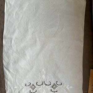 May include: A white linen towel with an embroidered decorative border. The border features a repeating pattern of openwork designs and floral elements. The towel is rectangular and appears to be folded. The fabric has a slightly textured appearance.
