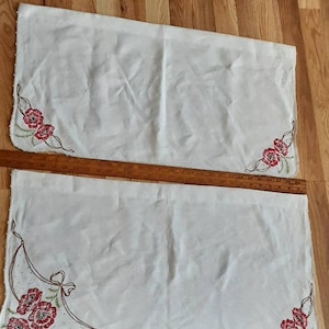 以下が含まれることがあります： 赤い花柄の刺繍が施された白いテーブルランナー2枚。ランナーは木製の床の上に置かれています。