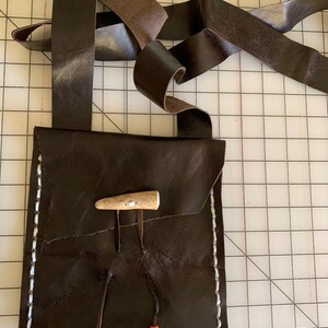 May include: A brown leather pouch with white stitching detail and a bone toggle closure. The pouch has two brown leather straps with red and black beads attached. The pouch is hanging from a brown leather strap.