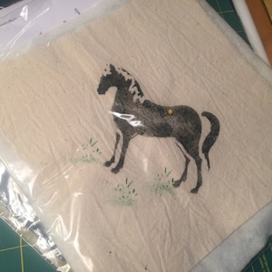 May include: A black and white horse silhouette stencil on a beige fabric background. The stencil is slightly faded and has a rustic look.