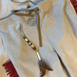 May include: Light gray pants with a drawstring and a decorative beaded clip featuring a feather. The pants have a leather drawstring and a small circular patch with text. Red fringe is visible on the side.
