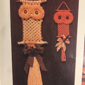 Puede incluir: Dos colgantes de pared de búho de macramé hechos a mano. El búho más grande es beige con ojos marrones y un lazo de lunares marrones y blancos. El búho más pequeño es naranja con ojos marrones y un lazo de estampado floral.