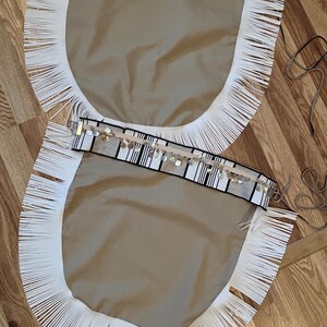 May include: Two beige fabric panels with white fringe trim. Each panel has a black and white striped border with silver embellishments. The panels are lying on a wooden surface.
