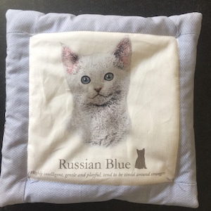 May include: A blue and white pillow with a picture of a Russian Blue cat on it. The text "Russian Blue" is printed below the cat, along with the description "Highly intelligent, gentle and playful, tend to be timid around strangers."