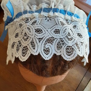 Puede incluir: Bonnet de encaje blanco para el cabello con un ribete de cinta azul. El bonnet se lleva en una cabeza de maniqu&iacute; con cabello casta&ntilde;o.