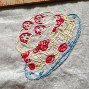 May include: A white fabric with a red and yellow embroidered design of a plate of spaghetti with tomato slices.