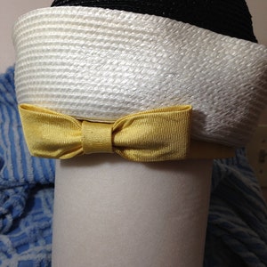 May include: A white and black straw hat with a yellow bow. The hat is sitting on a white surface.