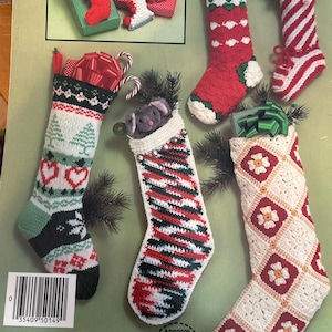 May include: A collection of handmade Christmas stockings in various designs. The stockings are red, white, green, and black, with patterns including stripes, trees, and hearts. Some stockings have candy canes and bows. The image is from the American School of Needlework.