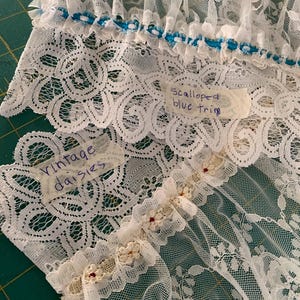 Puede incluir: Tela de encaje blanca con ribete azul festoneado y una etiqueta que dice "Scalloped blue trim". Otra etiqueta dice "Vintage daisies".