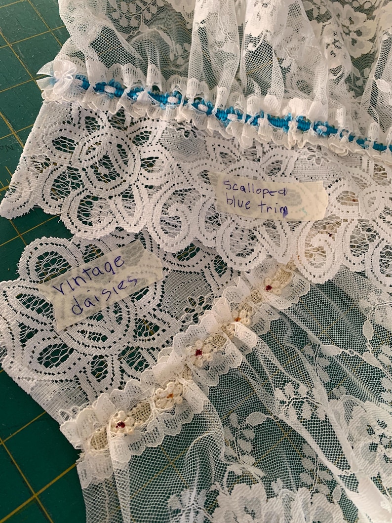 Puede incluir: Tela de encaje blanca con ribete azul festoneado y una etiqueta que dice "Scalloped blue trim". Otra etiqueta dice "Vintage daisies".