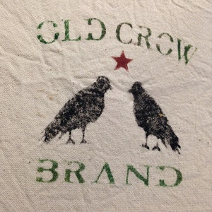 May include: A white fabric with a green and black stencil design. The stencil says "OLD CROW BRAND" with a red star and two black crow silhouettes.