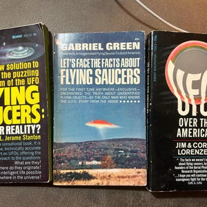 Op de afbeelding: Drie vintage paperback boeken over het onderwerp UFO's. De boekomslagen bevatten titels als "Flying Saucers" en "UFOs Over The Americas". De boeken liggen op een bruin oppervlak.