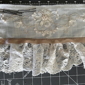May include: A piece of vintage fabric with a floral design and lace trim. The fabric is a light color, possibly off-white or cream, with embroidered flowers and a satin ribbon. Black bobby pins are visible.