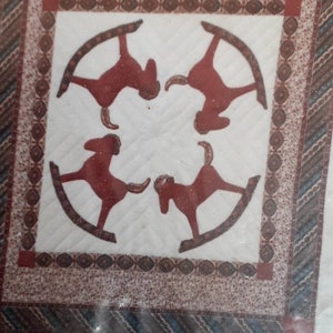 May include: A white quilted fabric with a brown and blue patterned border. The center of the fabric features a design of four red rocking horses in a circle.